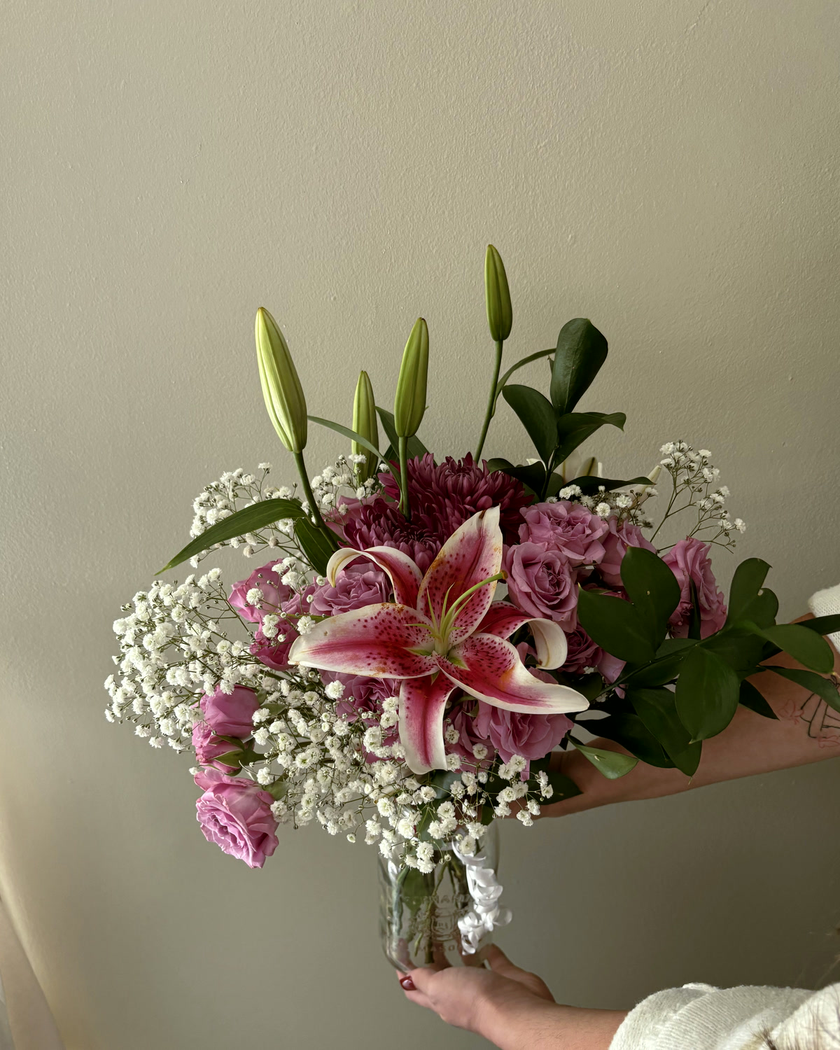 A finished bouquet of stargazer lily, pink roses and baby's breath held up against a plain wall