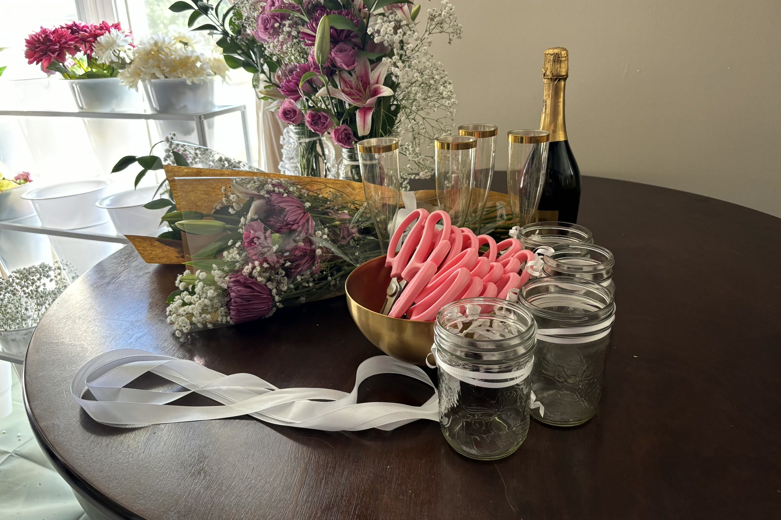 A bowl of pink snips, white ribbon, glass jars and a bottle of bubbly on the table